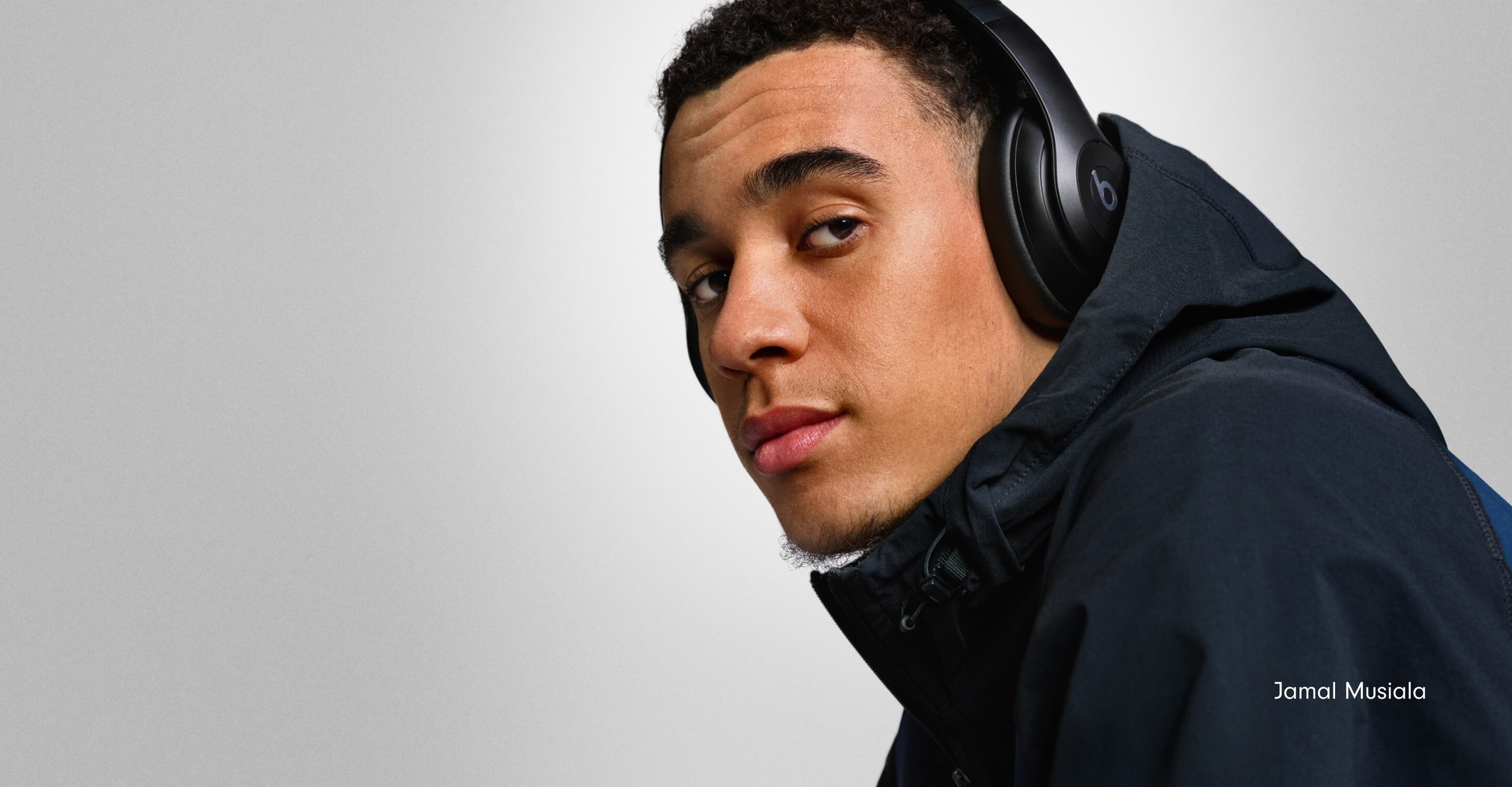Footballer Jamal Musiala wearing Beats Studio Pro headphones in Black against a light gray background, with the German text "Gestalte Deinen Spielraum" meaning "Design Your Playing Field."