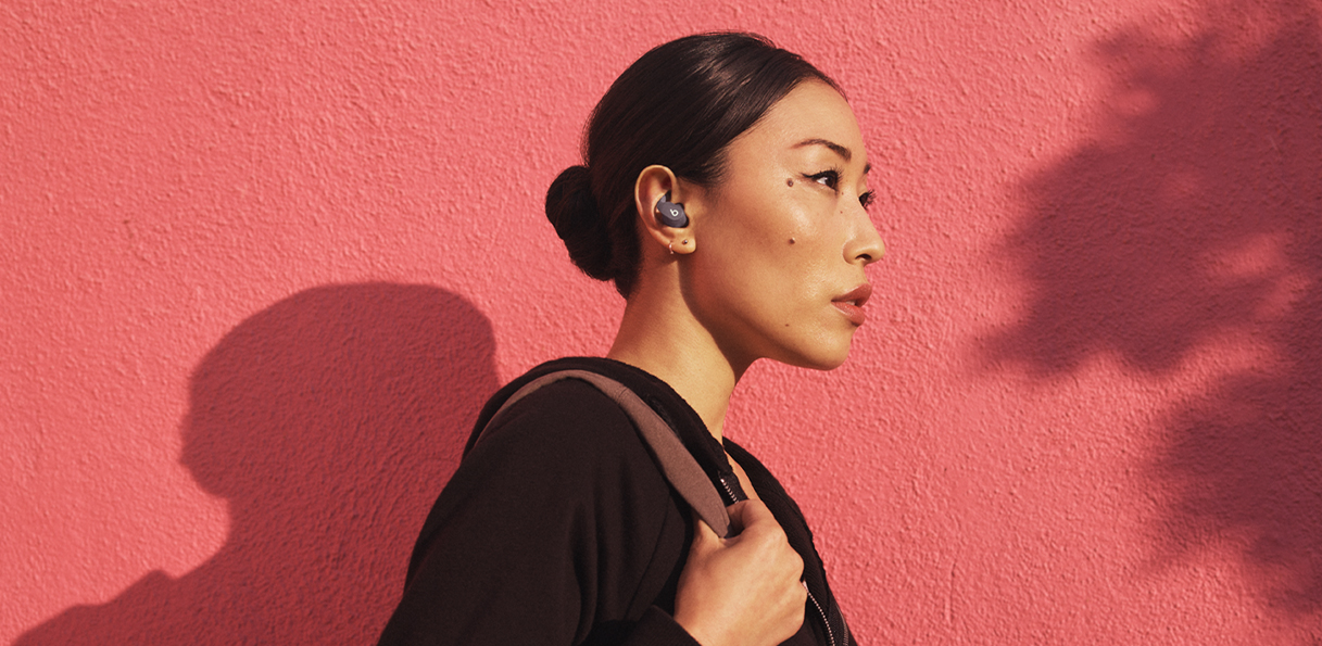 Asian woman wearing Beats Fit Pro while walking outside in front of a red wall