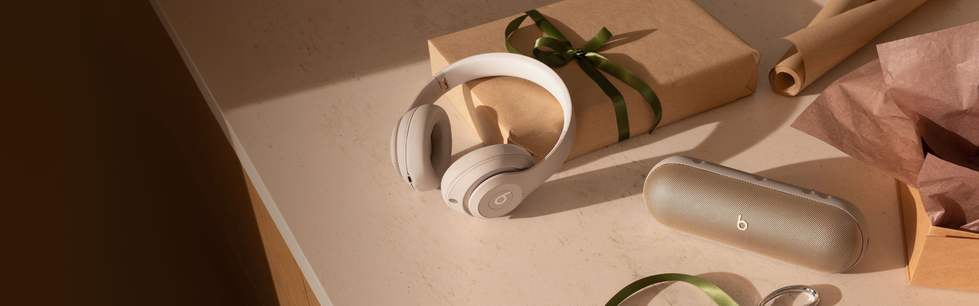 Beats Studio Pro in Sandstone and Beats Pill in Champagne Gold in front of wrapping paper and a gift on top of a counter.
