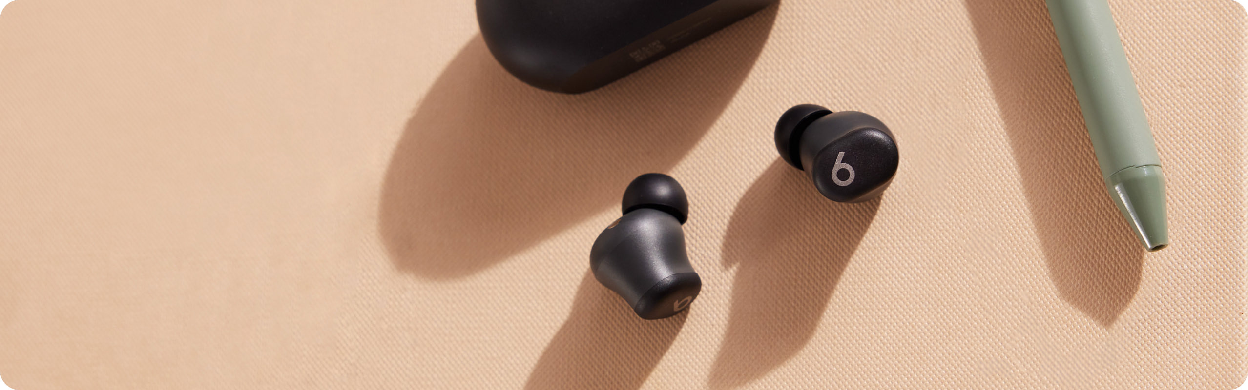 Two black Beats Solo Buds earbuds and their matching oval charging case are arranged on a light beige surface next to a green pen.